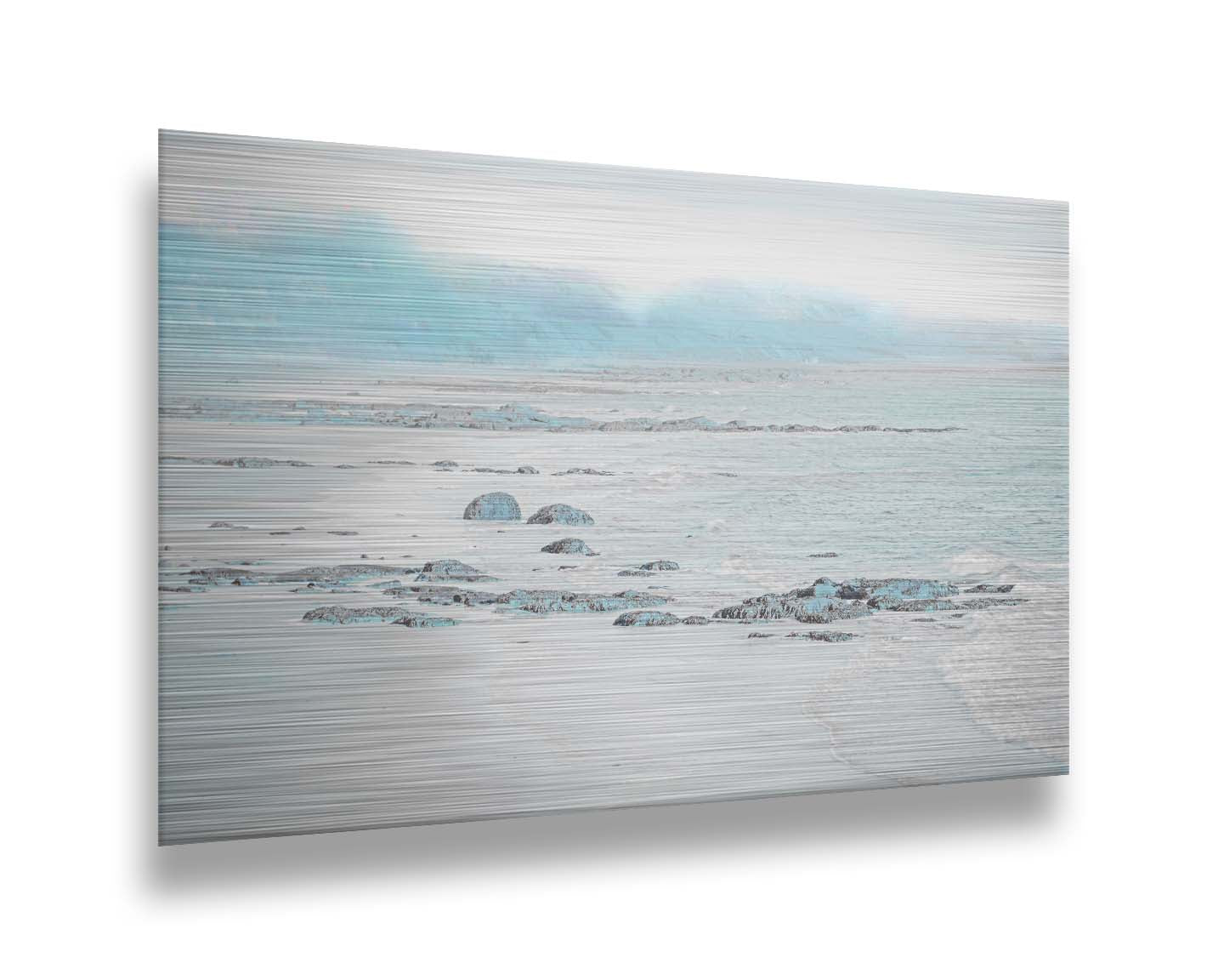 A photo of a rocky beach with a pale blue palette. The sea is calm and washing over the sand and rocks. Printed on metal.