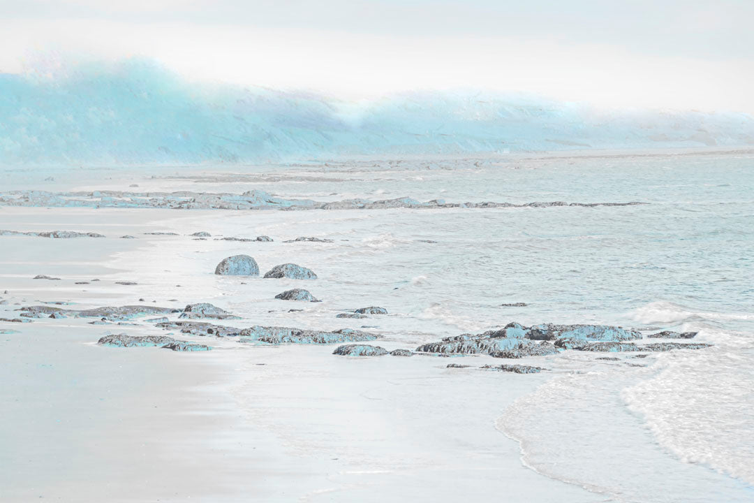 A photo of a rocky beach with a pale blue palette. The sea is calm and washing over the sand and rocks. 