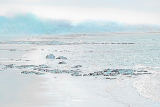 A photo of a rocky beach with a pale blue palette. The sea is calm and washing over the sand and rocks. 