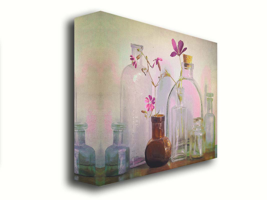 A photo of a selection of glass bottles and vases, sitting on a table. Sprigs of pink flowers rest in a few of the bottles. Printed on canvas.