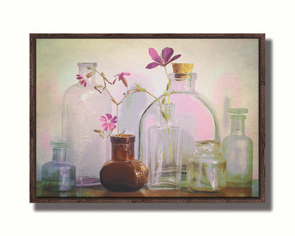 A photo of a selection of glass bottles and vases, sitting on a table. Sprigs of pink flowers rest in a few of the bottles. Printed on canvas in a float frame.