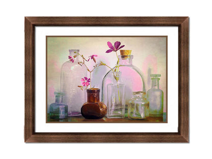 A photo of a selection of glass bottles and vases, sitting on a table. Sprigs of pink flowers rest in a few of the bottles. Printed on paper, matted, and framed.