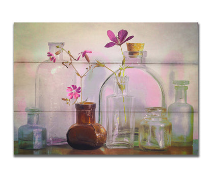 A photo of a selection of glass bottles and vases, sitting on a table. Sprigs of pink flowers rest in a few of the bottles. Printed on a box board.