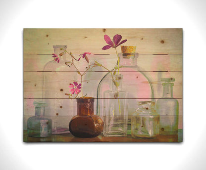 A photo of a selection of glass bottles and vases, sitting on a table. Sprigs of pink flowers rest in a few of the bottles. Printed on a wood pallet.