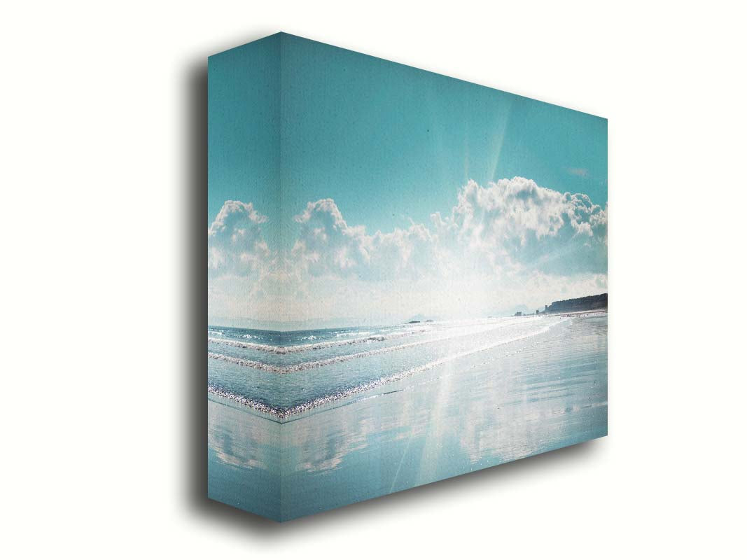 A photo of an ocean view, with a town on the coast. The sun is getting low in the sky, just before sunset, and the light is reflecting off the blue water and wet sand. Printed on canvas.