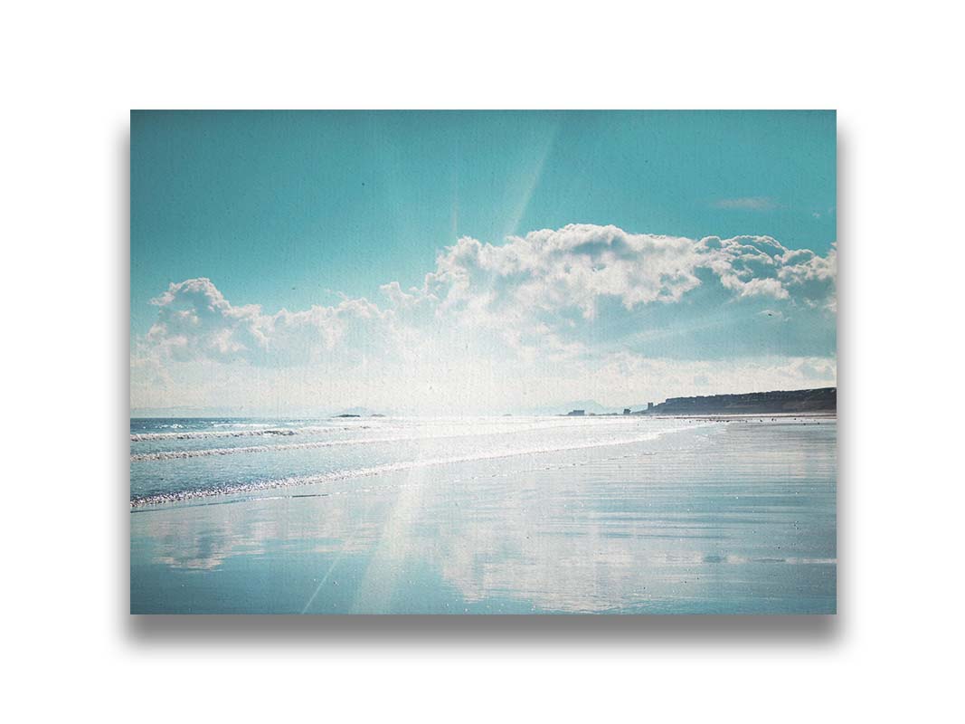 A photo of an ocean view, with a town on the coast. The sun is getting low in the sky, just before sunset, and the light is reflecting off the blue water and wet sand. Printed on canvas.