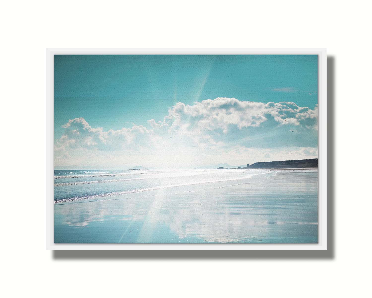 A photo of an ocean view, with a town on the coast. The sun is getting low in the sky, just before sunset, and the light is reflecting off the blue water and wet sand. Printed on canvas in a float frame.