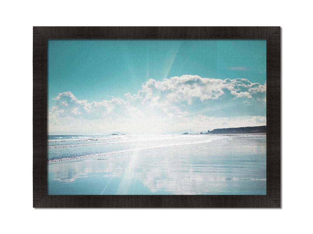 A photo of an ocean view, with a town on the coast. The sun is getting low in the sky, just before sunset, and the light is reflecting off the blue water and wet sand. Printed on canvas and framed.