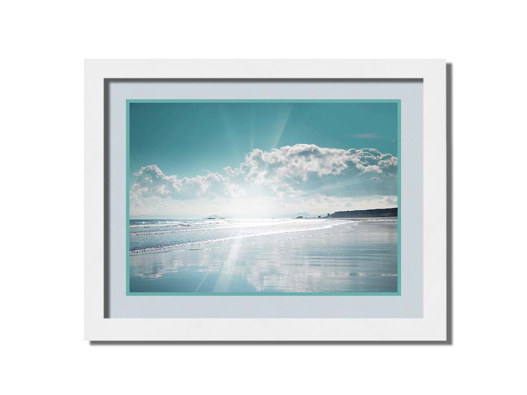 A photo of an ocean view, with a town on the coast. The sun is getting low in the sky, just before sunset, and the light is reflecting off the blue water and wet sand. Printed on paper, matted, and framed.