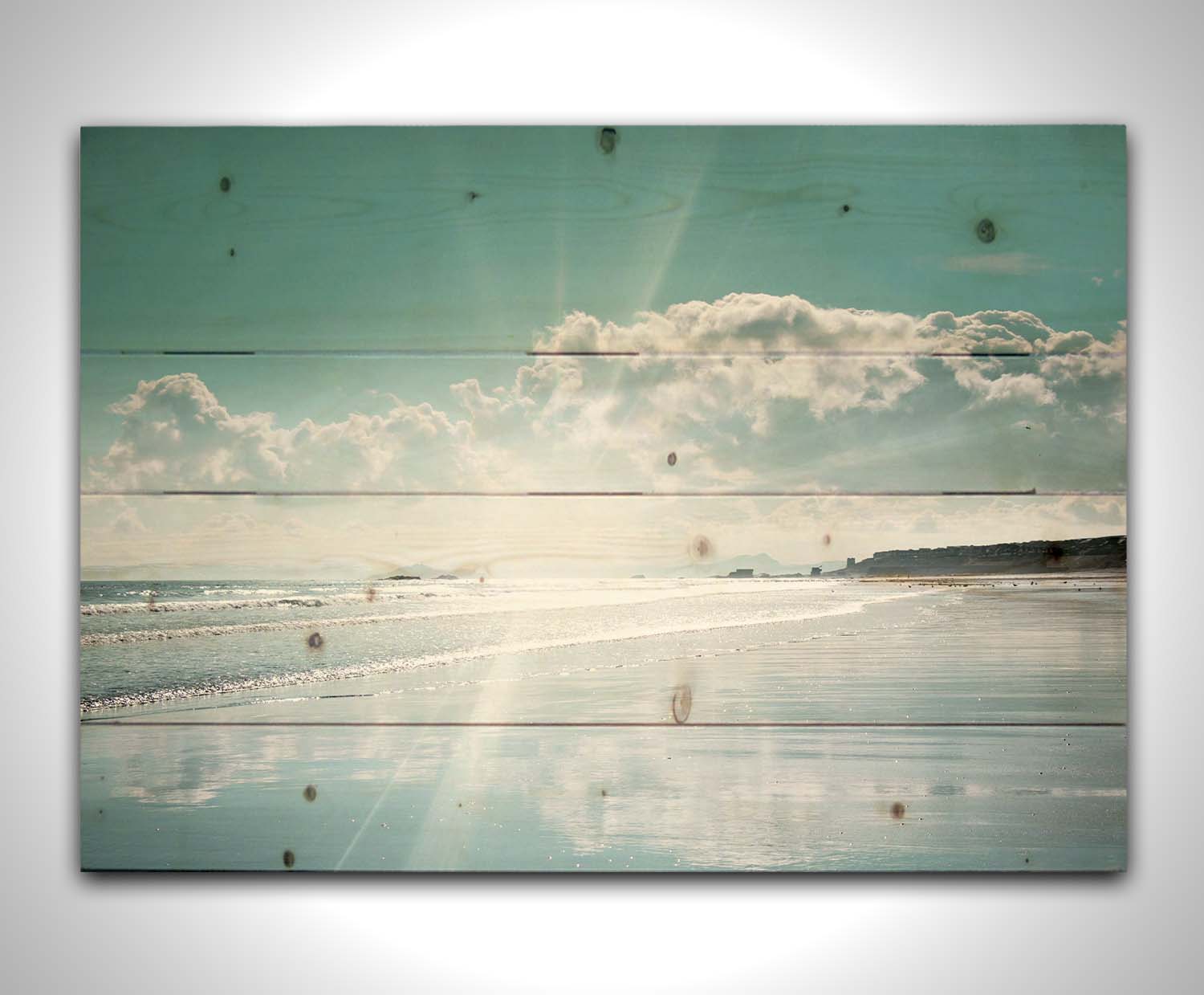 A photo of an ocean view, with a town on the coast. The sun is getting low in the sky, just before sunset, and the light is reflecting off the blue water and wet sand. Printed on a wood pallet.