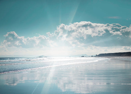 A photo of an ocean view, with a town on the coast. The sun is getting low in the sky, just before sunset, and the light is reflecting off the blue water and wet sand.
