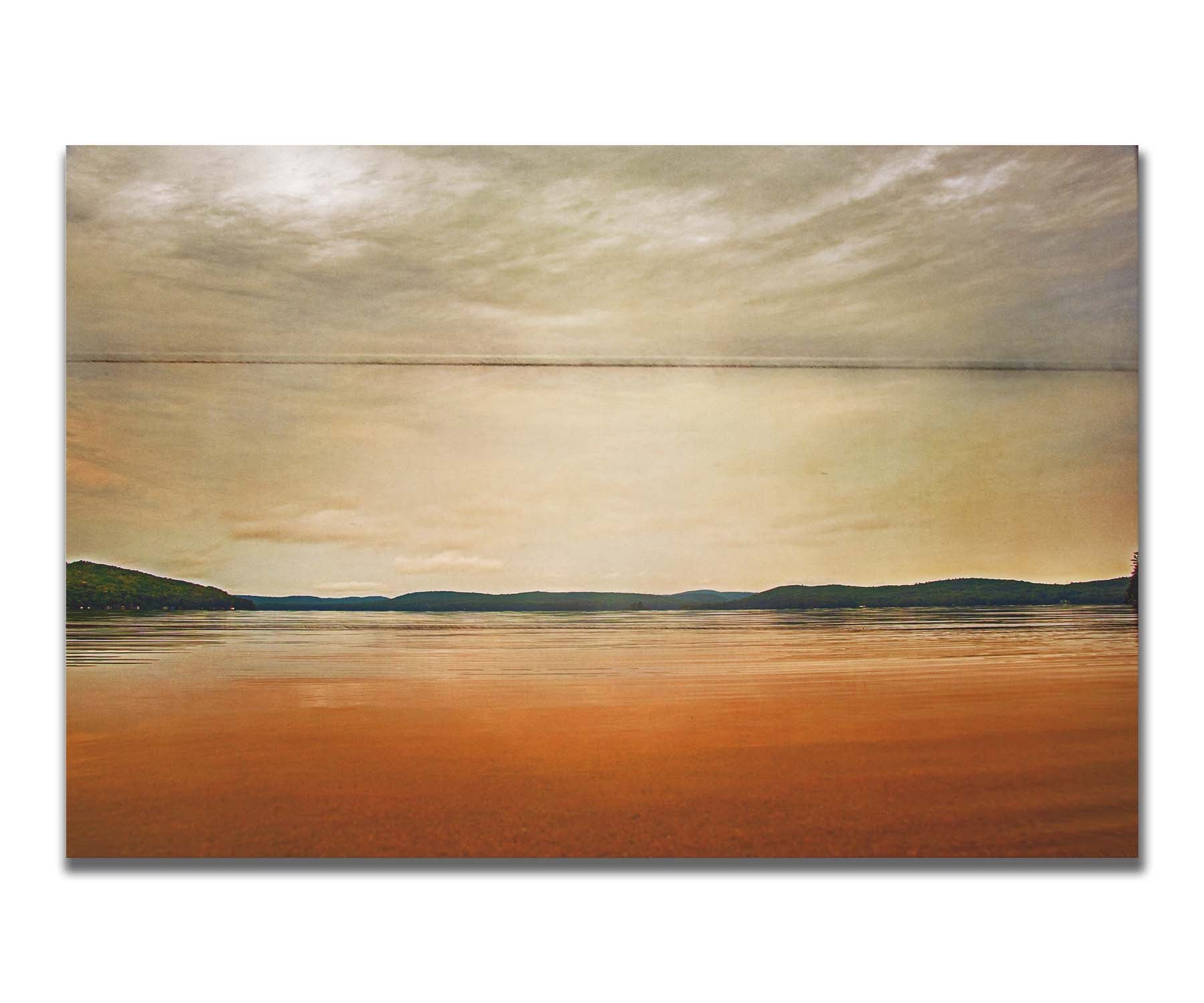 A photograph looking out at the sea and sky. The sky is cloudy and a warm gray, while the sea reflects a brilliant orange. Coastlines of trees can be seen in the distance. Printed on a box board.