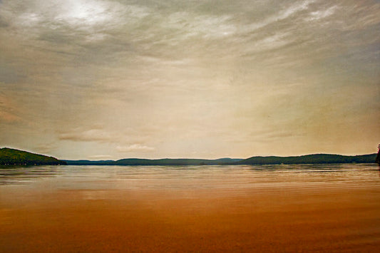 A photograph looking out at the sea and sky. The sky is cloudy and a warm gray, while the sea reflects a brilliant orange. Coastlines of trees can be seen in the distance. 
