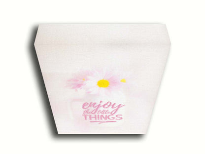 A photo of a white vase with pink daisies in a soft pink background, superimposed with the phrase "enjoy the little things". Printed on canvas.