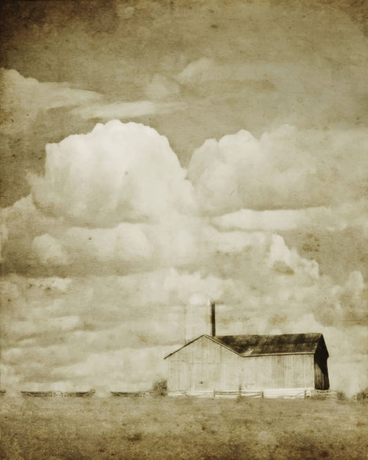 A sepia photo of a barn under a wide, open sky with large, fluffy clouds.