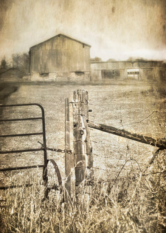 A photo of a farm fence and gate, with a large barn in the background in sepia tone. 