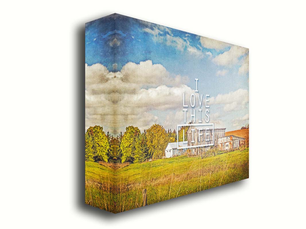 A photo of several barns, their associated pastures, and fields. The early signs of fall are present in the steadily changing leaves and crops. The sky above is wide, blue, and populated with fluffy clouds. It is overlaid with the phrase "I love this life." Printed on canvas.