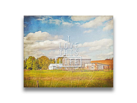 A photo of several barns, their associated pastures, and fields. The early signs of fall are present in the steadily changing leaves and crops. The sky above is wide, blue, and populated with fluffy clouds. It is overlaid with the phrase "I love this life." Printed on canvas.