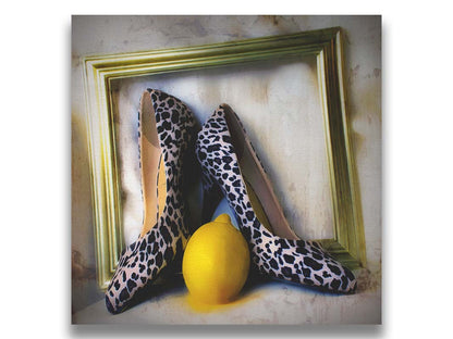 A still life photo of a pair of shoe heels in front of a gold picture frame, accompanied by a lemon. Printed on canvas.