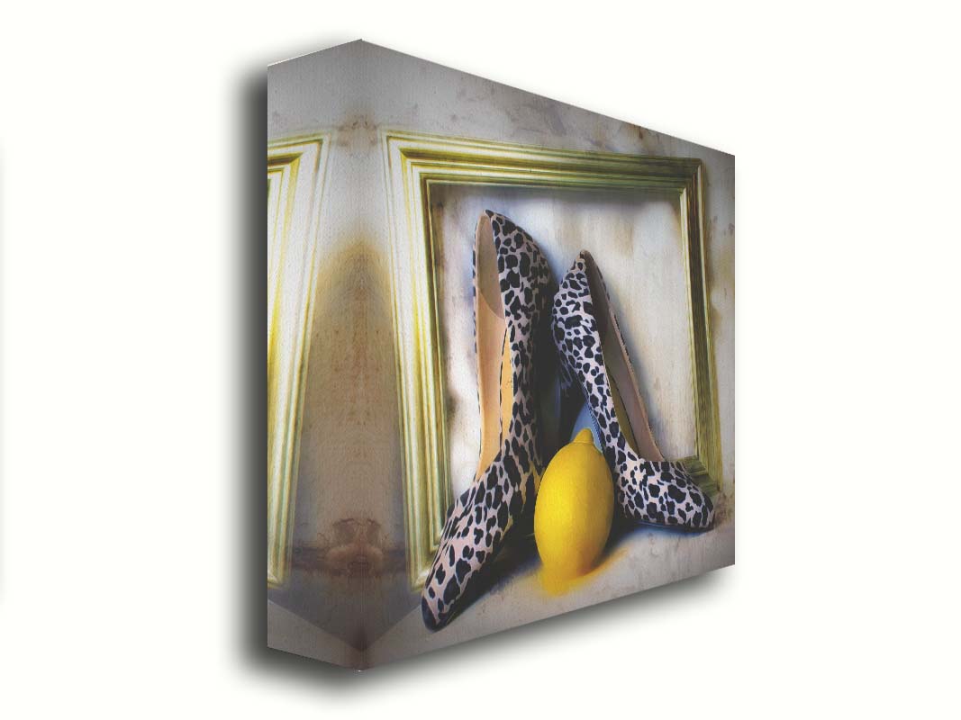 A still life photo of a pair of shoe heels in front of a gold picture frame, accompanied by a lemon. Printed on canvas.