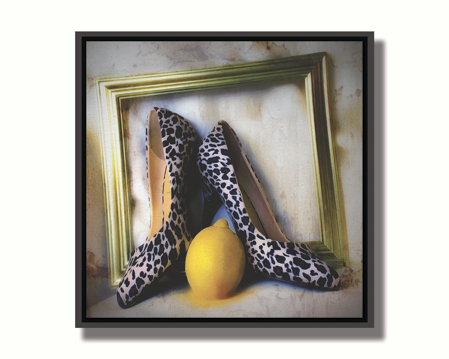 A still life photo of a pair of shoe heels in front of a gold picture frame, accompanied by a lemon. Printed on canvas in a float frame.