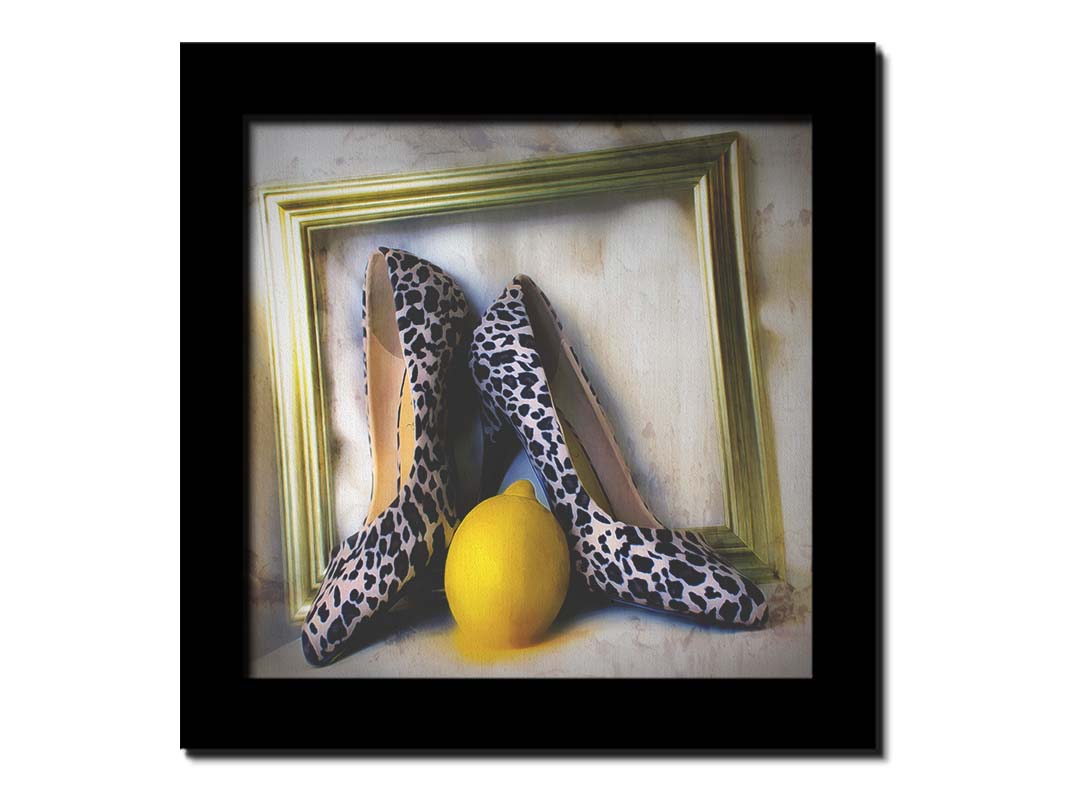 A still life photo of a pair of shoe heels in front of a gold picture frame, accompanied by a lemon. Printed on canvas and framed.