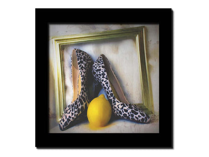 A still life photo of a pair of shoe heels in front of a gold picture frame, accompanied by a lemon. Printed on canvas and framed.