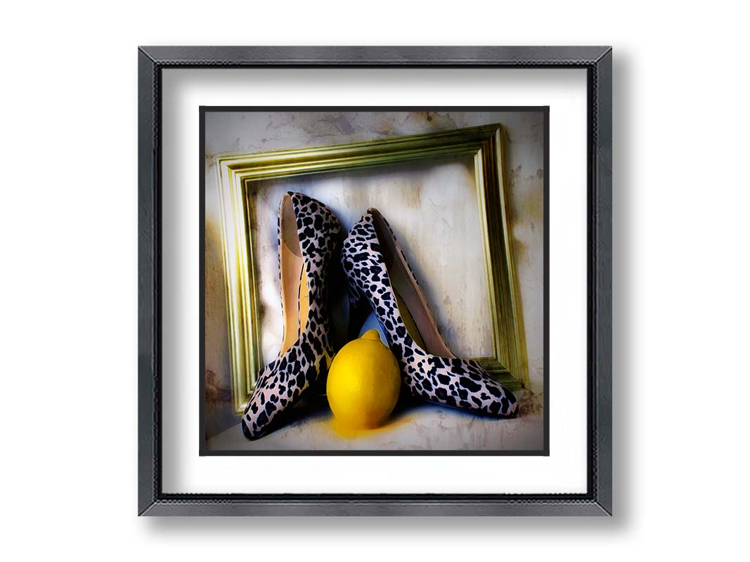 A still life photo of a pair of shoe heels in front of a gold picture frame, accompanied by a lemon. Printed on paper, matted, and framed.