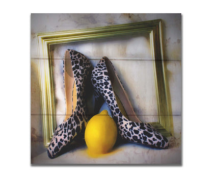 A still life photo of a pair of shoe heels in front of a gold picture frame, accompanied by a lemon. Printed on a box board.