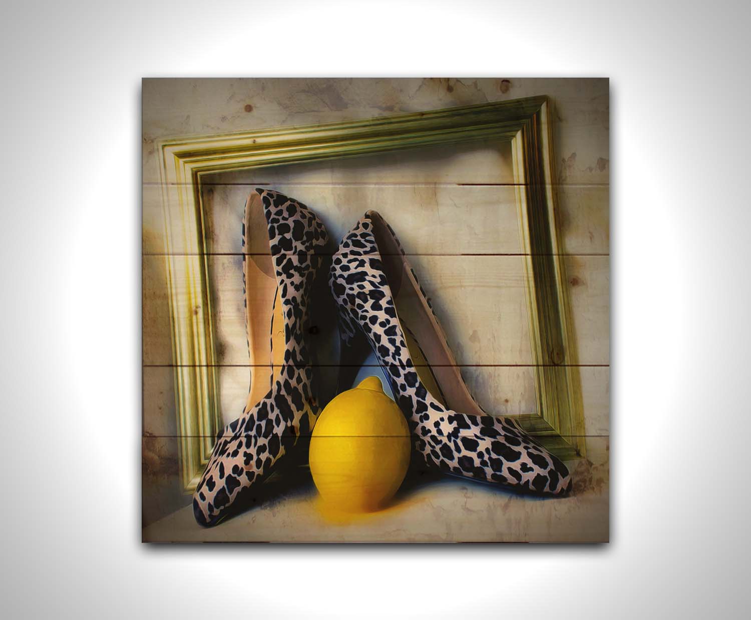 A still life photo of a pair of shoe heels in front of a gold picture frame, accompanied by a lemon. Printed on a wood pallet.