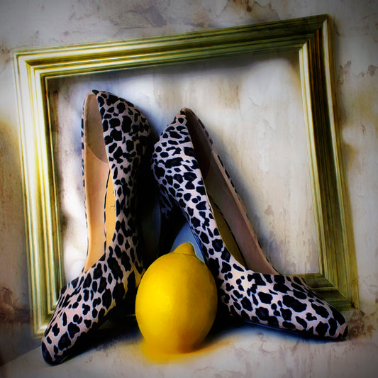 A still life photo of a pair of shoe heels in front of a gold picture frame, accompanied by a lemon.
