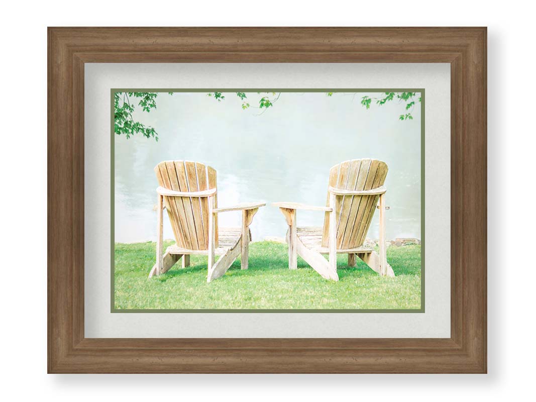 A photo of two wood lawn chairs, sitting on bright green grass, facing a lake. Printed on paper, matted, and framed.