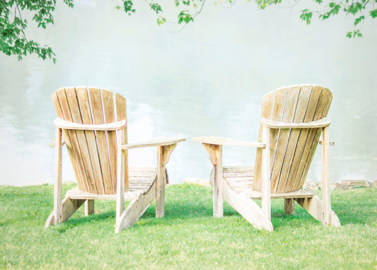 A photo of two wood lawn chairs, sitting on bright green grass, facing a lake. 