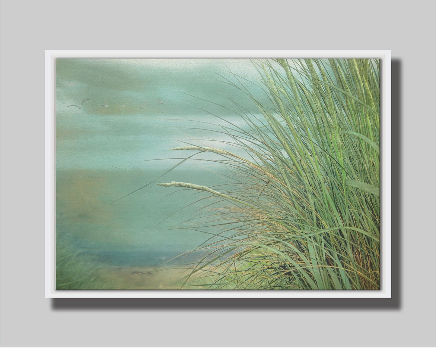 A photo of a tall grass plant on the beach, with the sea and cloudy skies in the background as seagulls fly above. Printed on canvas in a float frame.