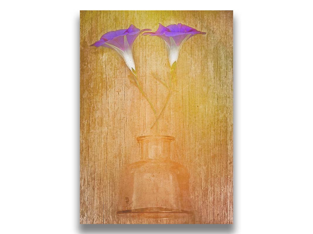 A photo of two purple flowers, crossed and lying on a yellow and orange wood background above a glass bottle. Printed on canvas.