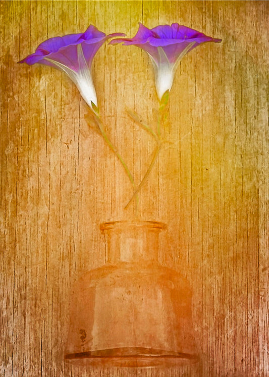 A photo of two purple flowers, crossed and lying on a yellow and orange wood background above a glass bottle. 