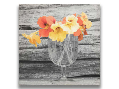 A grayscale photograph of a cup of water holding nasturtiums, against a wood wall. There is only one pop of color through the orange and yellow flower petals. Printed on canvas.