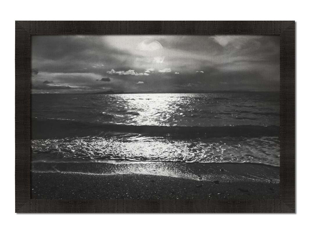 A photograph of the sea at night in grayscale. The light from the moon rerflects off the water in sharp contrast through the darkness of night. Printed on canvas and framed.