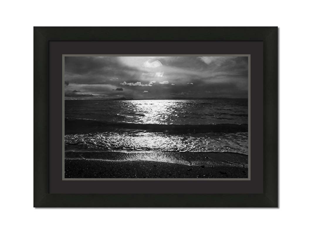 A photograph of the sea at night in grayscale. The light from the moon rerflects off the water in sharp contrast through the darkness of night. Printed on paper, matted, and framed.