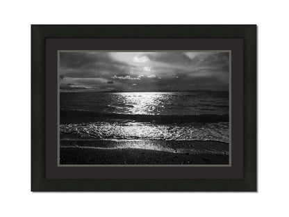 A photograph of the sea at night in grayscale. The light from the moon rerflects off the water in sharp contrast through the darkness of night. Printed on paper, matted, and framed.