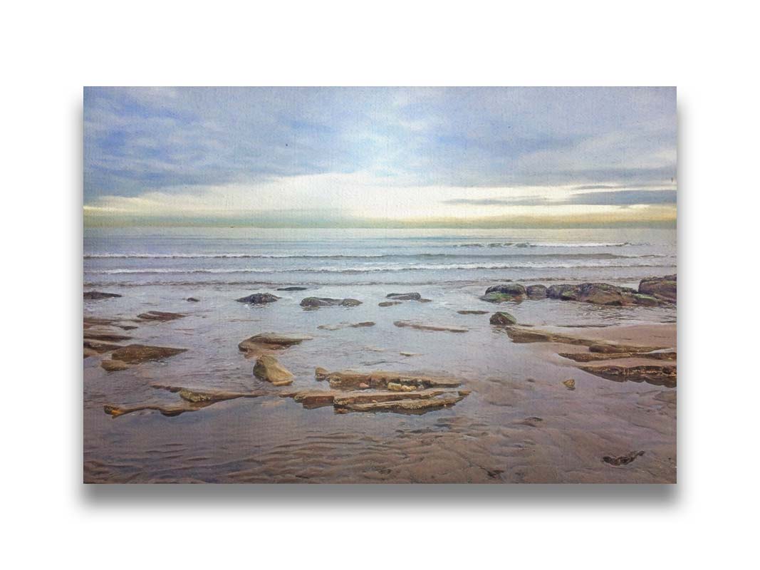 A photograph of the rocky shore of the North Sea at low tide. The sun peeks over the horizon through the partly cloudy sky. Printed on canvas.