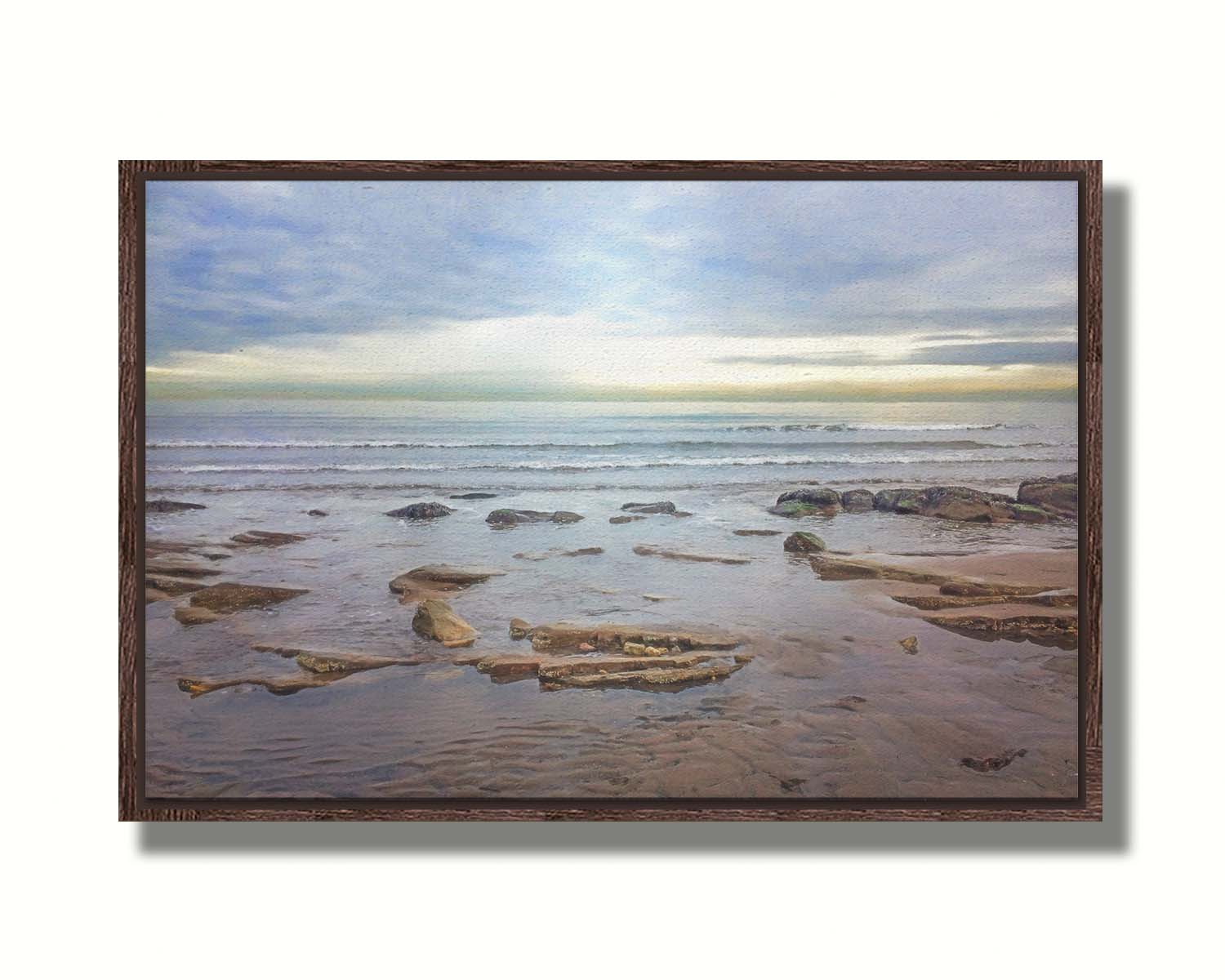 A photograph of the rocky shore of the North Sea at low tide. The sun peeks over the horizon through the partly cloudy sky. Printed on canvas in a float frame.