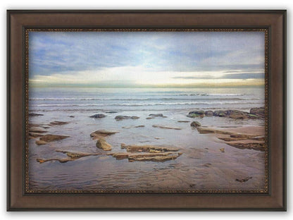 A photograph of the rocky shore of the North Sea at low tide. The sun peeks over the horizon through the partly cloudy sky. Printed on canvas and framed.