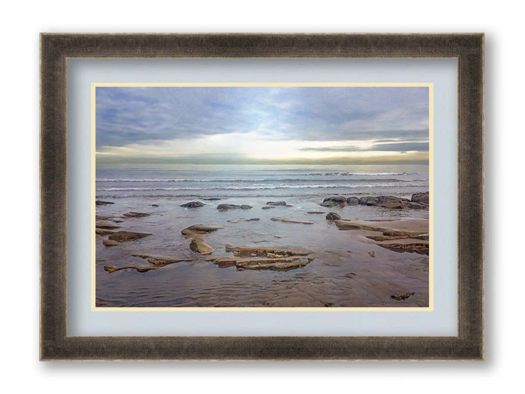 A photograph of the rocky shore of the North Sea at low tide. The sun peeks over the horizon through the partly cloudy sky. Printed on paper, matted, and framed.