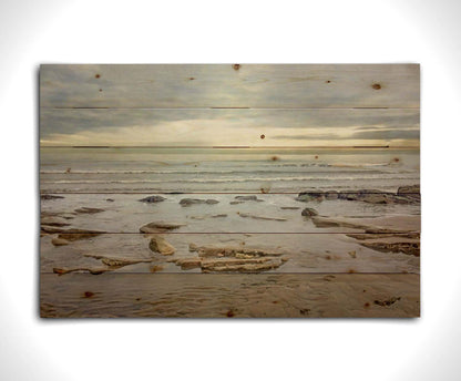 A photograph of the rocky shore of the North Sea at low tide. The sun peeks over the horizon through the partly cloudy sky. Printed on a wood pallet.