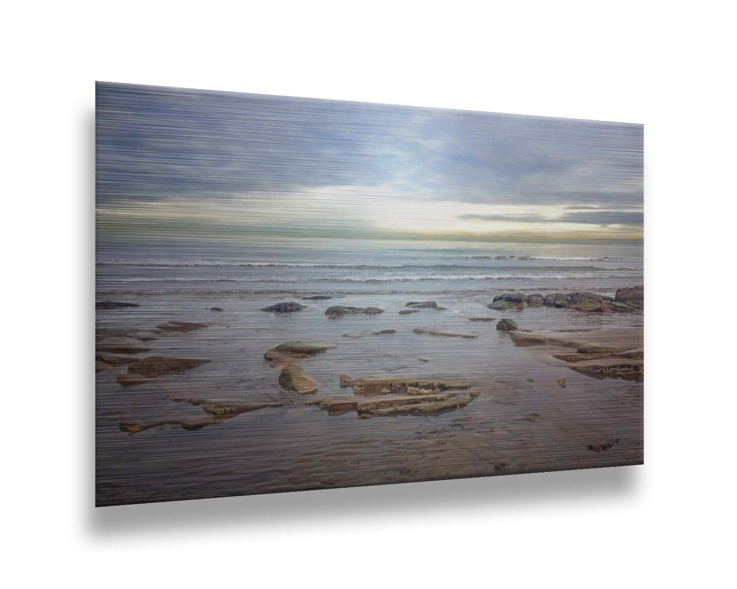 A photograph of the rocky shore of the North Sea at low tide. The sun peeks over the horizon through the partly cloudy sky. Printed on metal.