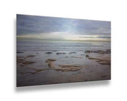 A photograph of the rocky shore of the North Sea at low tide. The sun peeks over the horizon through the partly cloudy sky. Printed on metal.