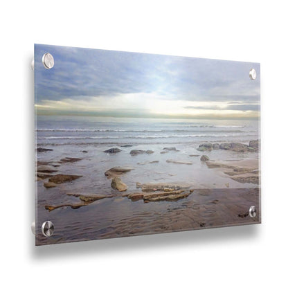 A photograph of the rocky shore of the North Sea at low tide. The sun peeks over the horizon through the partly cloudy sky. Printed on acrylic.