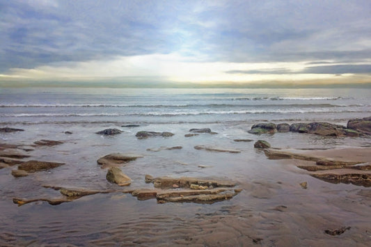 A photograph of the rocky shore of the North Sea at low tide. The sun peeks over the horizon through the partly cloudy sky.