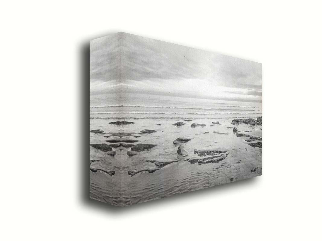 A grayscale photograph of the rocky shore of the North Sea at low tide. The sun peeks over the horizon through the partly cloudy sky. Printed on canvas.